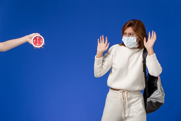 Vooraanzicht jonge vrouwelijke student in witte trui met masker bang voor vrouw met klok op blauwe muur