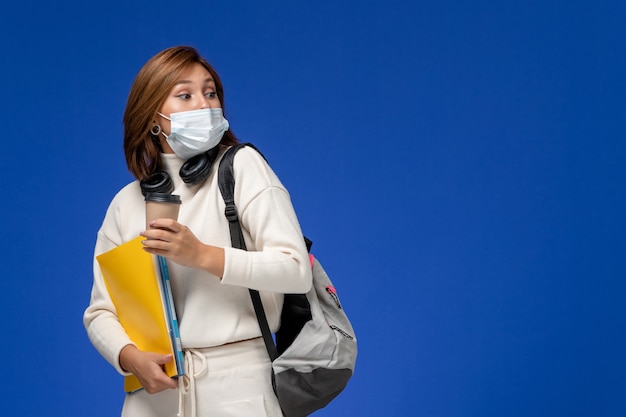 Vooraanzicht jonge vrouwelijke student in witte trui met masker en rugzak met bestanden en koffie op het blauwe bureau college universiteit boek lessen