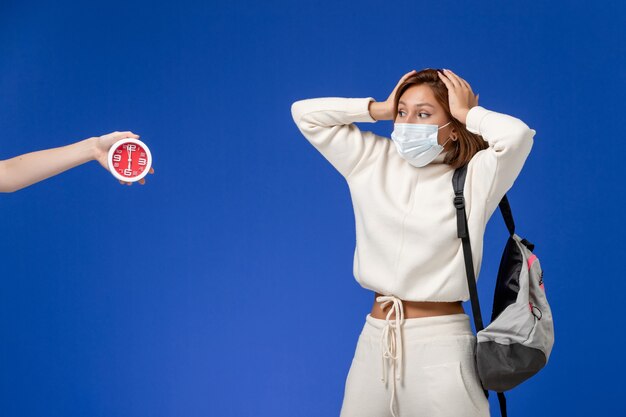 Vooraanzicht jonge vrouwelijke student in witte trui met masker en tas op de blauwe muur