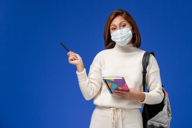 Vooraanzicht jonge vrouwelijke student in witte trui met masker met tas en voorbeeldenboek met pen op blauwe muur