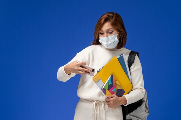 Vooraanzicht jonge vrouwelijke student in witte trui met masker met tas en voorbeeldenboeken en met behulp van haar telefoon op de blauwe muur