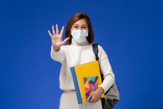 Vooraanzicht jonge vrouwelijke student in witte trui met masker met tas en voorbeeldenboeken op blauw bureau lessen college universiteit school meisje