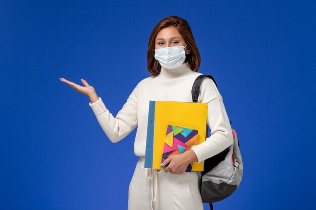 Vooraanzicht jonge vrouwelijke student in witte trui met masker met tas en voorbeeldenboeken op de blauwe muur