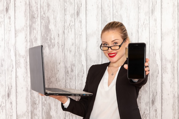 Vooraanzicht jonge zakenvrouw in strikte kleren zwarte jas met behulp van haar laptop en telefoon glimlachend op wit oppervlak te houden