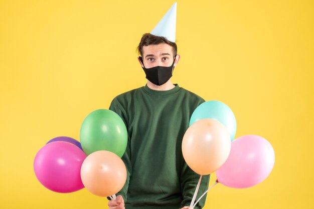 Vooraanzicht jongeman met feestmuts met kleurrijke ballonnen staande op gele achtergrond