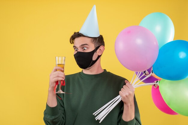 Vooraanzicht jongeman met grote ogen met feestmuts en zwart masker met wijnglas en ballonnen op geel