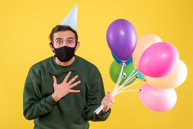 Vooraanzicht jongeman met grote ogen met feestmuts hand op zijn borst zetten staande op geel