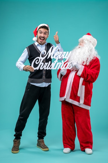 Gratis foto vooraanzicht kerstman met jonge mannelijke bedrijf vrolijk kerstfeest schrijven op de blauwe achtergrond