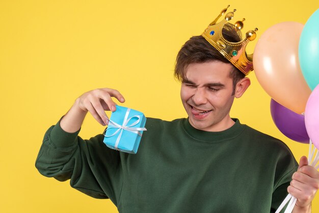 Vooraanzicht knappe jongeman met kroon met ballonnen en cadeau op geel