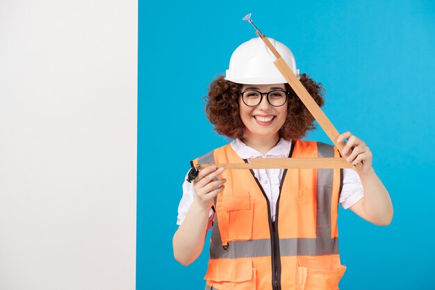 Vooraanzicht lachende vrouwelijke bouwer in uniform met houten gereedschap op blauw