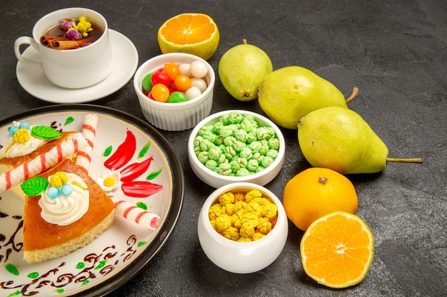 Gratis foto vooraanzicht lekkere slagroomtaart cake plakjes met kopje thee peren en snoepjes op donkere ruimte