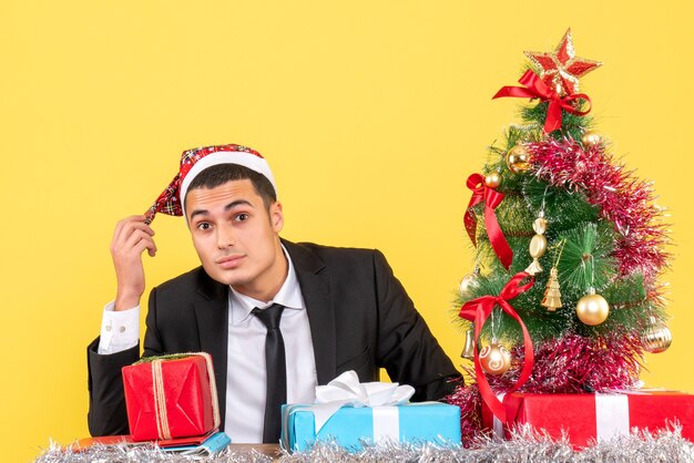 Vooraanzicht man in pak met kerstmuts zittend aan de tafel kerstboom en geschenken