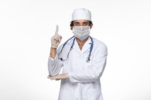 Vooraanzicht mannelijke arts in medisch pak die een steriel masker draagt als een bescherming tegen covid - op witte muur virusziekte covid pandemie