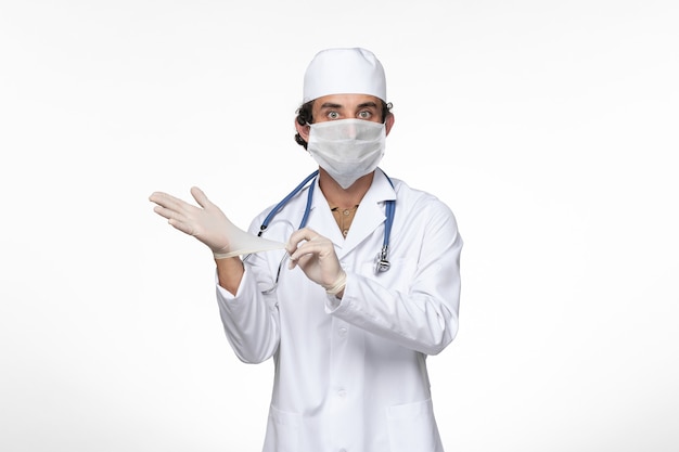 Vooraanzicht mannelijke arts in medisch pak die steriel masker draagt als bescherming tegen covid - handschoenen dragen op wit bureau virusziekte covid pandemische ziekte