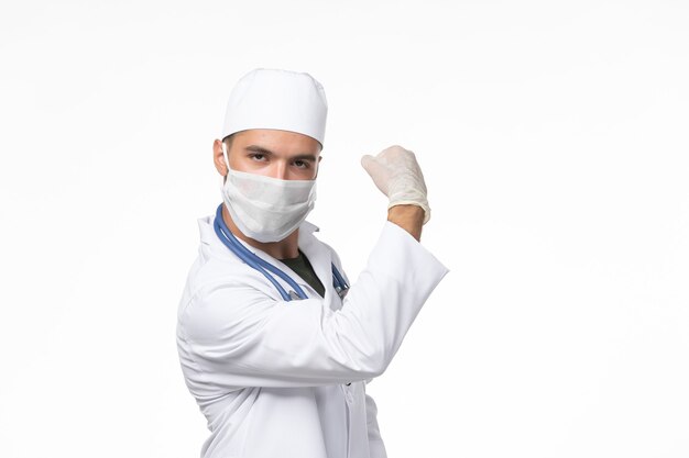 Vooraanzicht mannelijke arts in medisch pak en draagt masker vanwege covid- op witte muur covid-virus geneeskunde pandemische ziekte