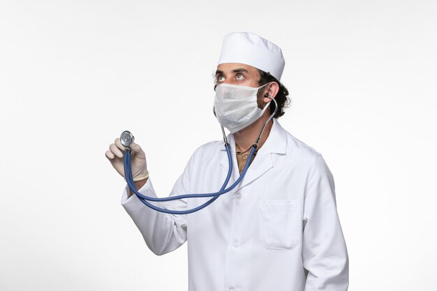 Vooraanzicht mannelijke arts in medisch pak en het dragen van een masker als bescherming tegen covid - met behulp van stethoscoop op witte muur ziekte virus covid - pandemie