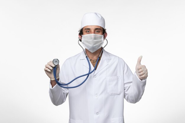 Vooraanzicht mannelijke arts in medisch pak en het dragen van een masker als een bescherming tegen covid - met behulp van stethoscoop op wit bureau ziekte virus covid - pandemie