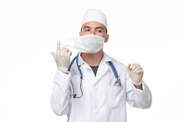 Vooraanzicht mannelijke arts in medisch pak en het dragen van masker vanwege covid-on the light wall ziekte virus covid-pandemische ziekte