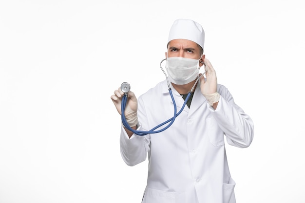Vooraanzicht mannelijke arts in medisch pak en masker tegen coronavirus met behulp van een stethoscoop op de witte muur covid- ziekte ziekte pandemie