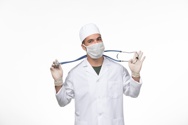 Vooraanzicht mannelijke arts in medisch pak en masker tegen coronavirus met stethoscoop op witte muur covid-virus ziekte ziekte pandemie