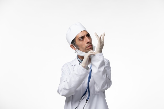 Vooraanzicht mannelijke arts in medisch pak en masker vanwege coronavirus handschoenen dragen op een wit oppervlak