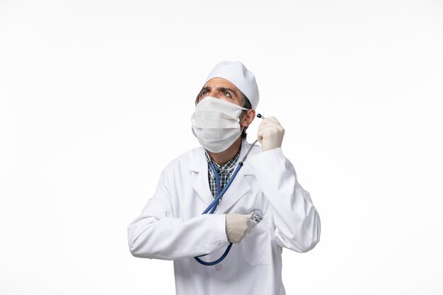 Vooraanzicht mannelijke arts in medisch pak en masker vanwege coronavirus met behulp van een stethoscoop op wit bureau