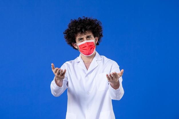 Vooraanzicht mannelijke arts in medisch pak met rood masker op blauwe achtergrond medicijnvirus covid-vaccin gezondheid geneeskunde ziekenhuispandemie