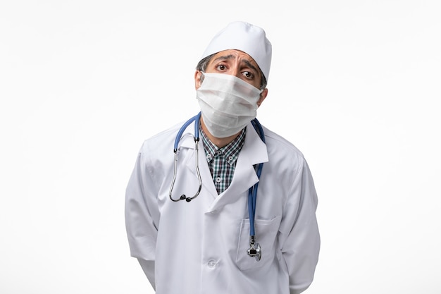Vooraanzicht mannelijke arts in wit medisch pak met masker vanwege coronavirus poseren op wit oppervlak