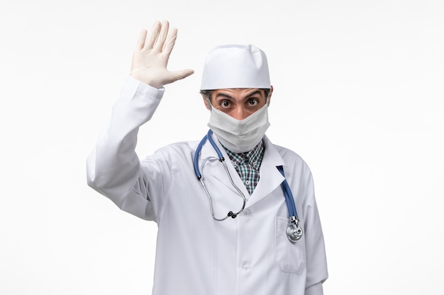 Vooraanzicht mannelijke arts in wit medisch pak met masker vanwege covid - op witte vloer ziekte virus covid - ziekenhuis pandemie