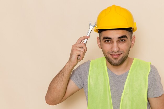 Gratis foto vooraanzicht mannelijke bouwer in gele helm die zilveren hulpmiddel op de lichte achtergrond houdt