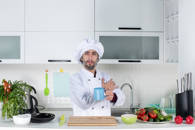 Vooraanzicht mannelijke chef-kok in koksmuts met beker die achter de keukentafel staat