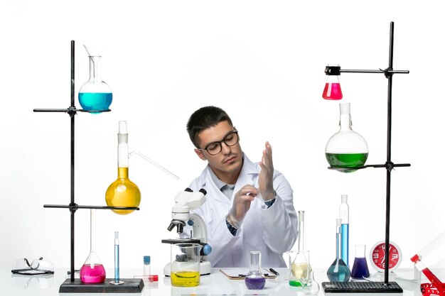 Vooraanzicht mannelijke chemicus in witte medische pak zitten met oplossingen handschoenen dragen op witte achtergrond virus lab covid ziekte wetenschap
