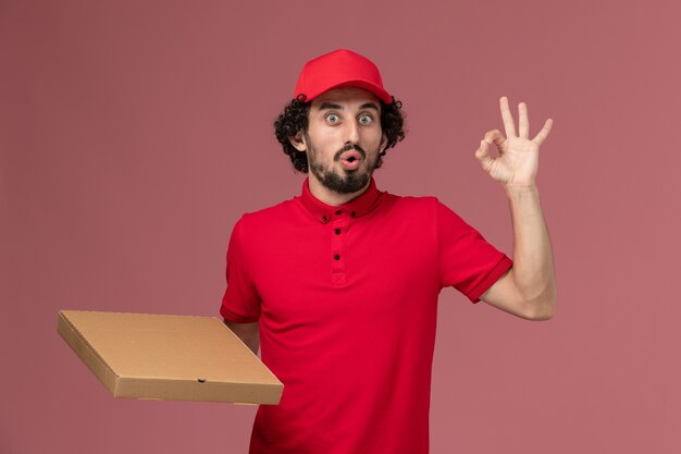 Vooraanzicht mannelijke koerier bezorger in rood shirt en cape bedrijf levering voedsel doos op roze muur service levering bedrijf werknemer werk baan