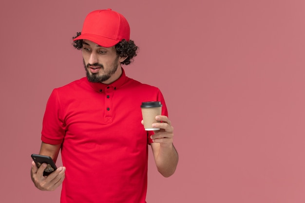 Vooraanzicht mannelijke koerier bezorger in rood shirt en cape met bruine bezorging koffiekopje en telefoon op lichtroze muur service bezorgmedewerker werknemer baan