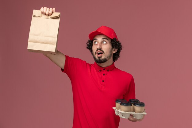 Vooraanzicht mannelijke koerier bezorger in rood shirt en cape met bruine bezorging koffiekopjes met voedselpakket op roze muur servicebedrijf werker