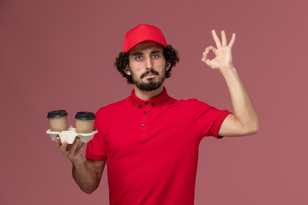 Vooraanzicht mannelijke koerier bezorger in rood shirt en cape met bruine bezorging koffiekopjes op lichtroze muur service baan bezorgmedewerker