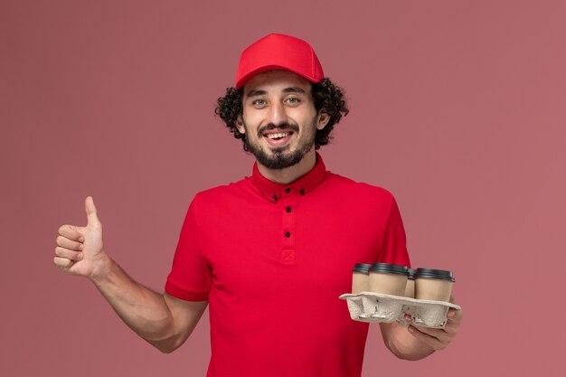 Vooraanzicht mannelijke koerier bezorger in rood shirt en cape met bruine bezorging koffiekopjes op lichtroze muur servicewerknemer