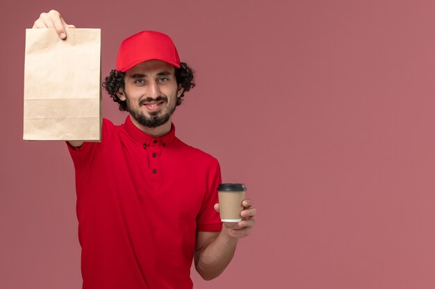 Vooraanzicht mannelijke koerier bezorger in rood shirt en cape met bruine koffiekopje en voedselpakket op lichtroze muur service bezorgwerk werknemer baan