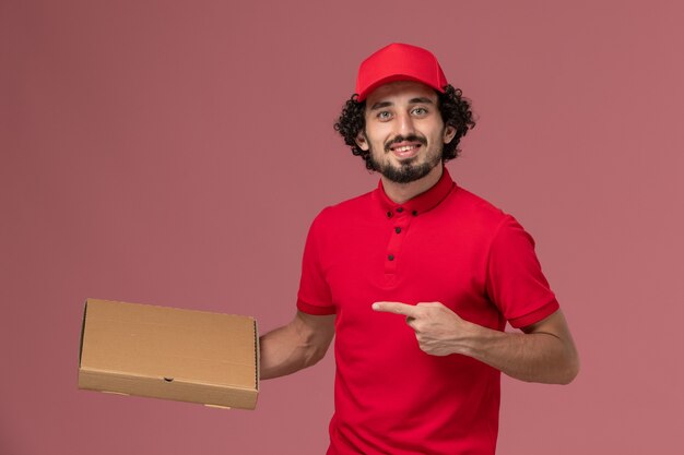 Vooraanzicht mannelijke koerier bezorger in rood shirt en cape met levering voedsel doos op roze muur service bezorgbedrijf werknemer werk