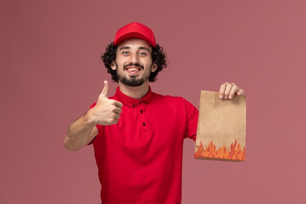 Vooraanzicht mannelijke koerier bezorger in rood shirt en cape met voedselpakket en lachend op roze muur service bezorgbedrijf werknemer