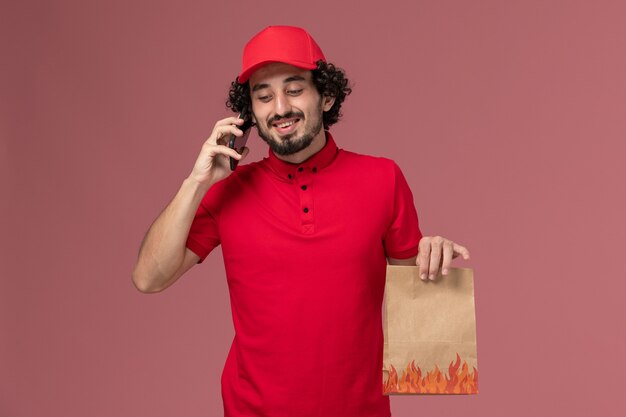 Vooraanzicht mannelijke koerier bezorger in rood shirt en cape met voedselpakket praten aan de telefoon op roze muur service levering werknemer baan