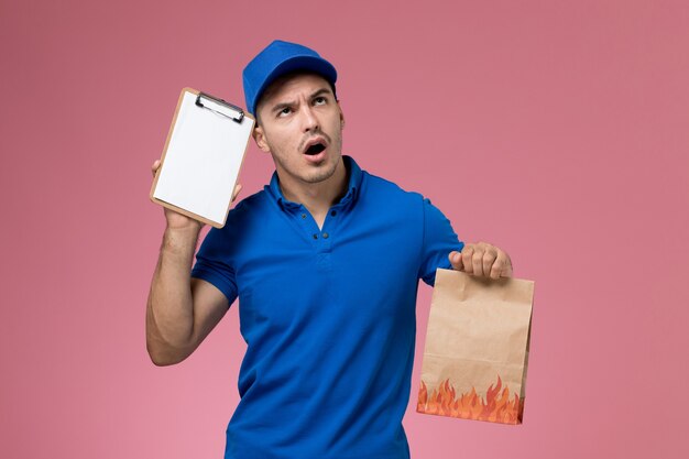 Vooraanzicht mannelijke koerier in blauw uniform bedrijf blocnote papier pakket op roze muur, werknemer uniforme dienstverlening