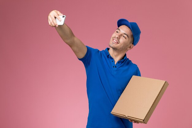 Vooraanzicht mannelijke koerier in blauw uniform die een foto neemt met voedseldoos op roze muur, levering van de arbeidersuniforme dienst