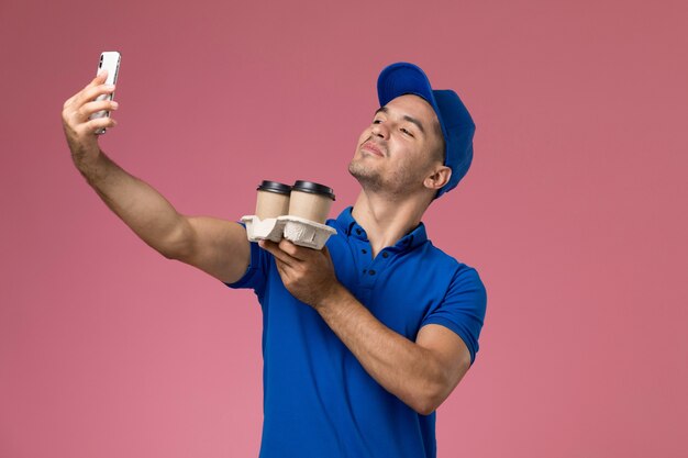 Vooraanzicht mannelijke koerier in blauw uniform die een selfie met koffiekopjes op roze muur, werknemer uniforme dienstverlening