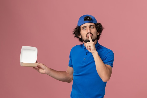 Vooraanzicht mannelijke koerier in blauw uniform en cape die een leeg klein bezorgvoedselpakket op het roze muur uniforme bezorgbedrijf houdt