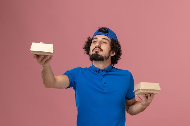 Vooraanzicht mannelijke koerier in blauw uniform en cape die kleine voedselpakketjes op de roze muur houdt