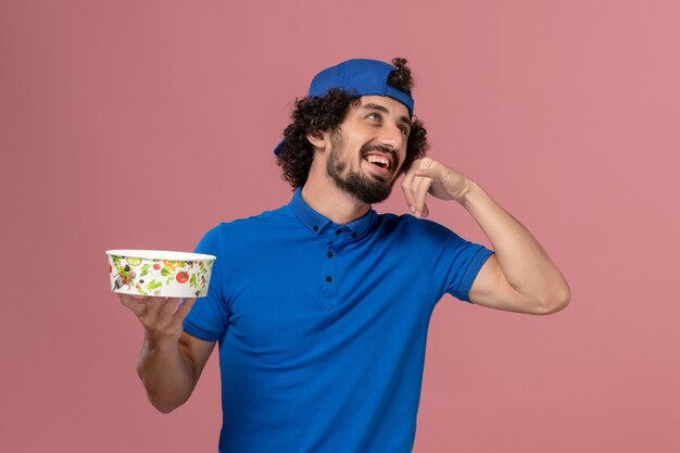 Gratis foto vooraanzicht mannelijke koerier in blauw uniform en cape die ronde leveringskom op de roze muur houden