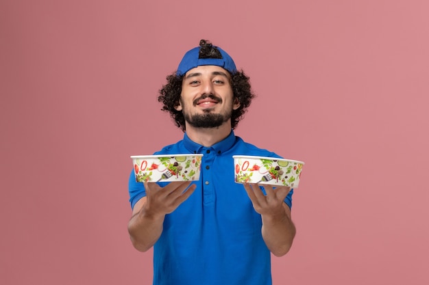 Gratis foto vooraanzicht mannelijke koerier in blauw uniform en cape die ronde leveringskommen op de roze muur houden