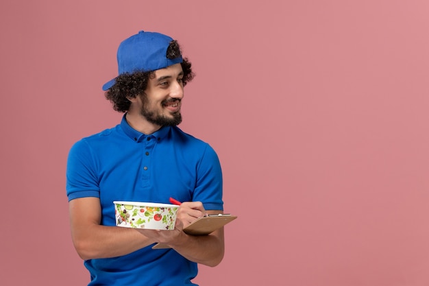 Vooraanzicht mannelijke koerier in blauw uniform en cape met blocnote en ronde leveringskom die op de roze muur schrijft