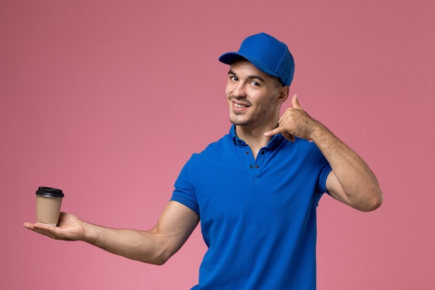 Vooraanzicht mannelijke koerier in blauw uniform glimlachend en levering koffiekopje op de roze muur, uniforme dienstverlening baan levering te houden